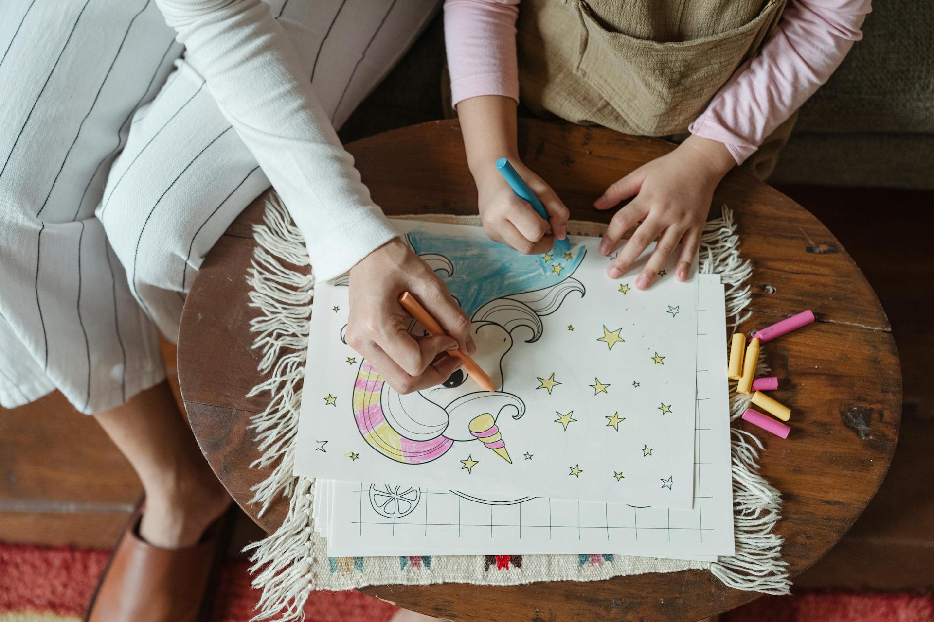 From above of crop unrecognizable mother and daughter using wax pencils for coloring drawing with unicorn on coffee table at home