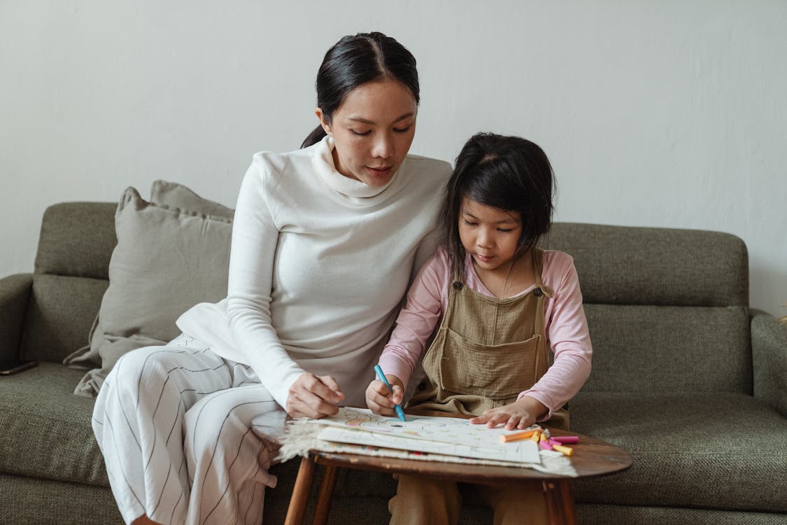 Free Concentrated little Asian girl in casual clothes studiously drawing picture on paper while mother watching process sitting together on sofa Stock Photo