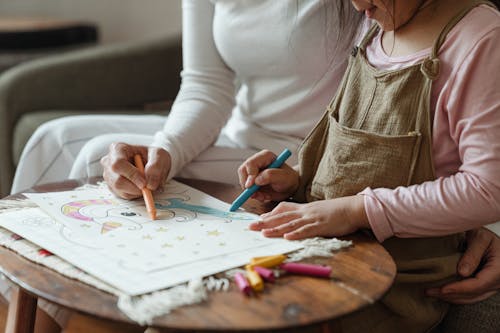 Crop mother and daughter coloring drawing together on coffee table