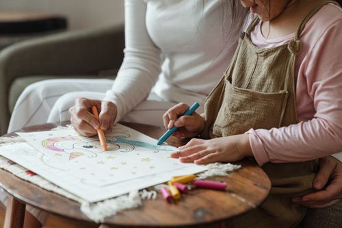 Free Side view of crop anonymous woman and little girl in casual clothes sitting on sofa and coloring drawing with wax colored pencils on coffee table Stock Photo
