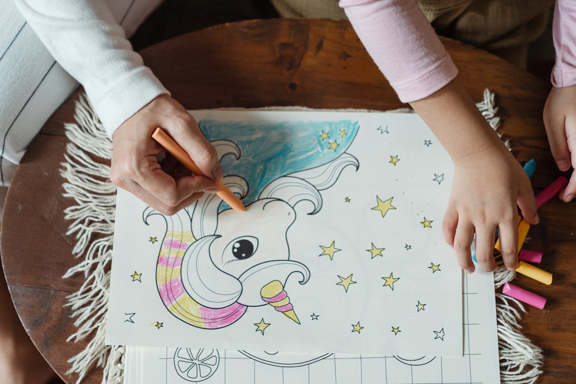 Free From above of crop anonymous mother and daughter coloring picture with unicorn with wax crayons on coffee table Stock Photo