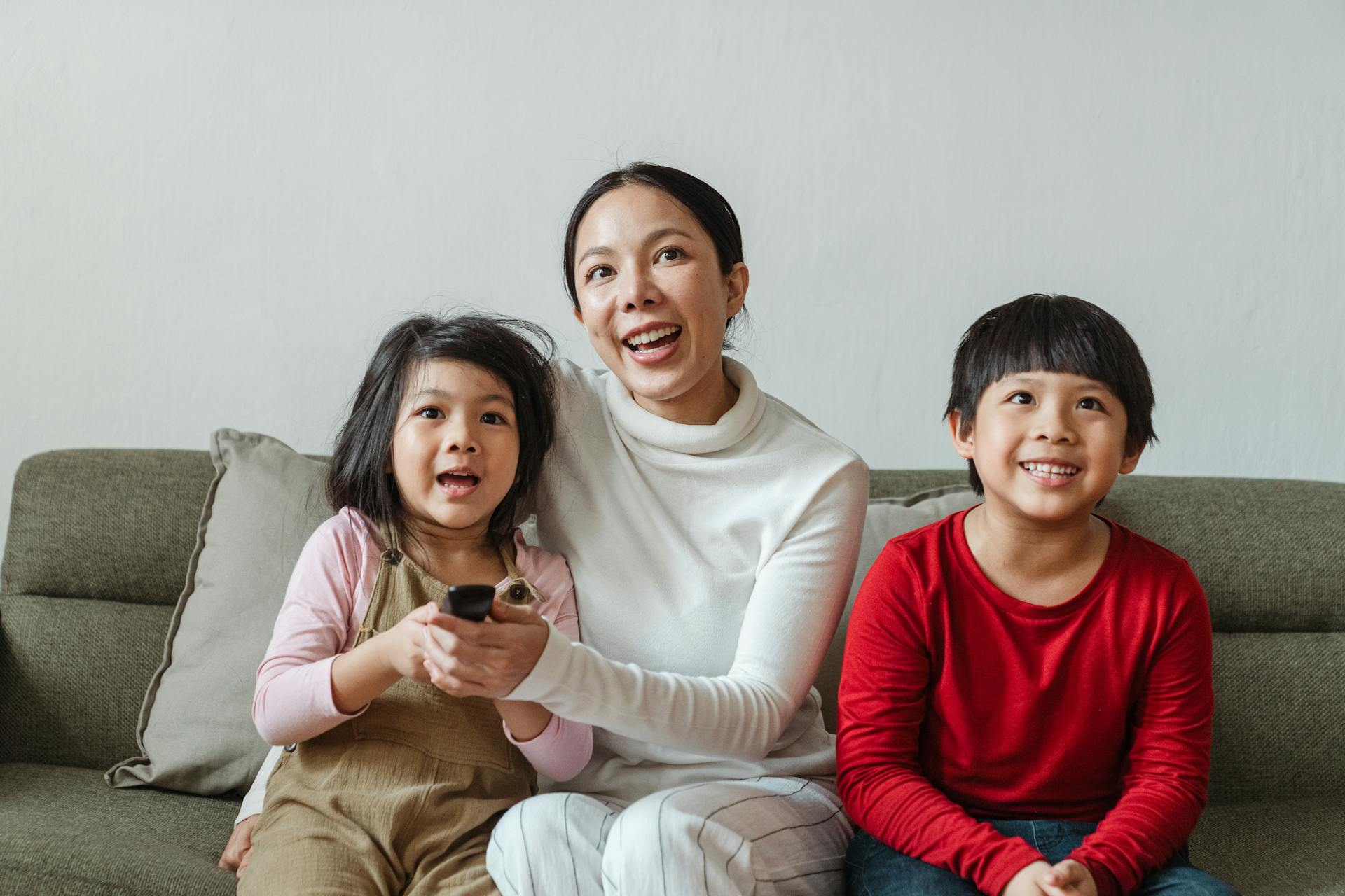 Optimistic Asian woman with little daughter and son in casual outfit sitting on sofa while choosing television channel using remote