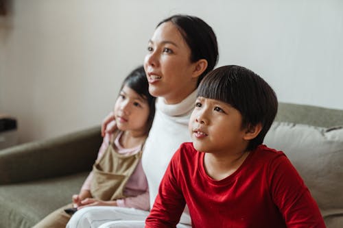 Focused Asian little boy watching movie on TV together with mother embracing sister while having pastime in living room in daytime