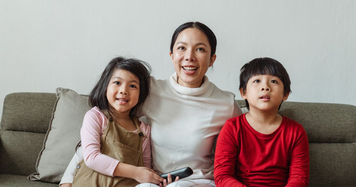 Positive Asian mother hugging little son and daughter sitting on sofa while watching funny movie on TV channel together
