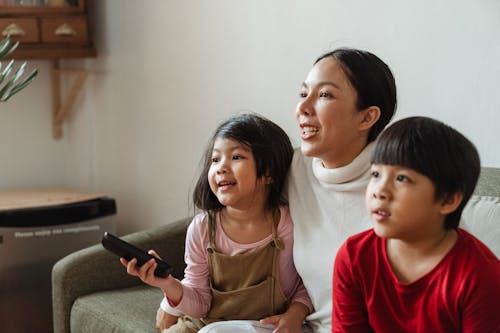 Happy Asian mother hugging little cute children on couch and watching entertaining program on TV