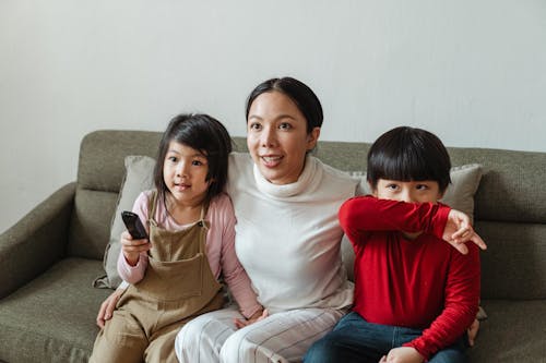 Concentrated young ethnic woman cuddling little children while watching interesting video together on comfortable couch