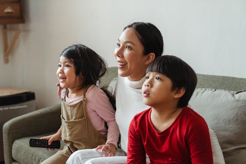 Cute little Asian kids watching exciting movie with mom while resting on sofa at home