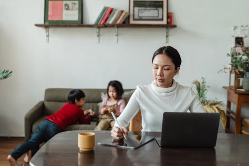 Focused young Asian mother working remotely while children playing on sofa