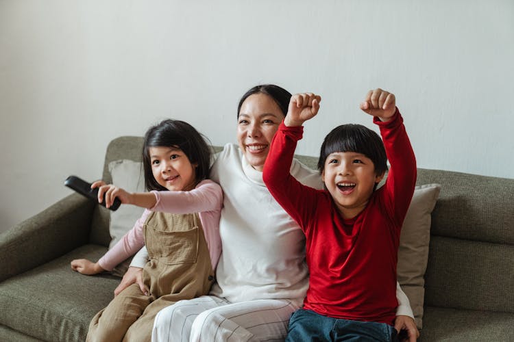 Cute Ethnic Little Kids Cheering While Watching TV On Sofa With Mom