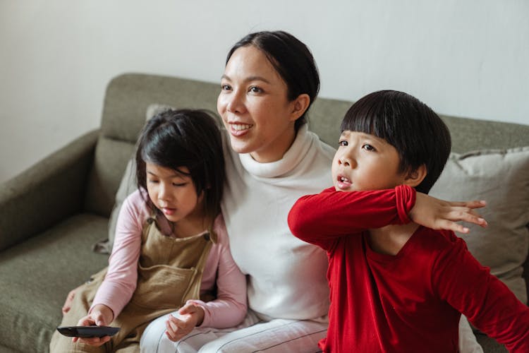 Cute Ethnic Kids And Mother Sitting On Sofa And Watching TV With Attention