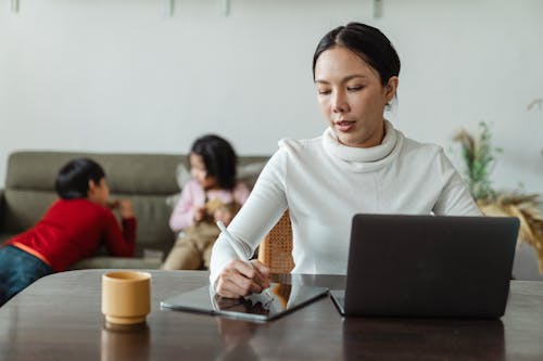 Focused young Asian woman in casual wear writing with stylus on tablet and browsing laptop while working remotely at home with children