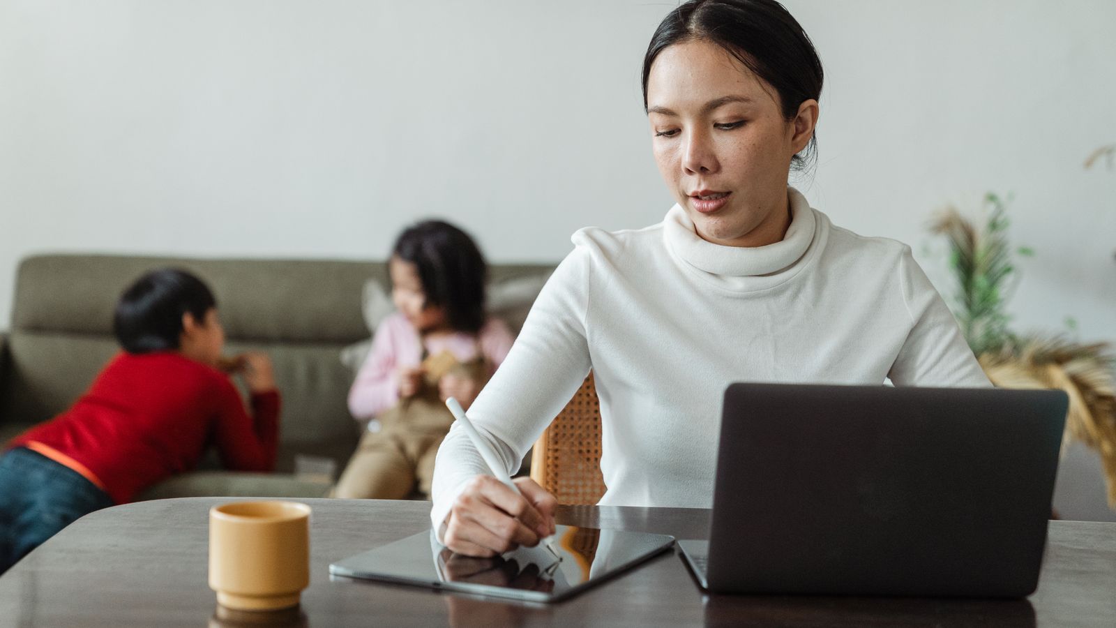 Smiling mom using a laptop at the kitchen table while her toddler plays beside her
