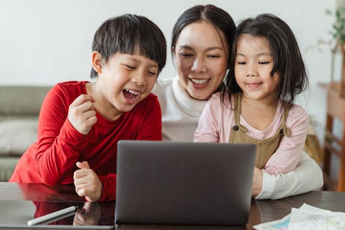 Cheerful little Asian boy and girl watching funny video on laptop with happy smiling mother during weekend