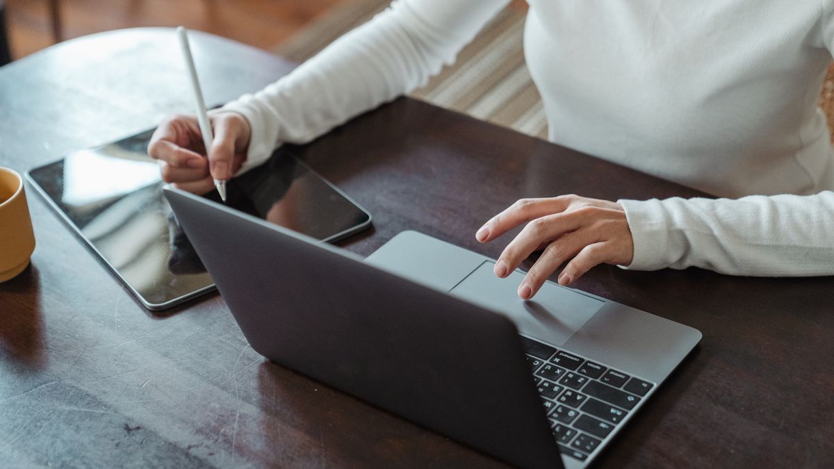 From above of crop anonymous businesswoman in white sweater working at table using tablet with stylus and typing on laptop