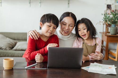 Positive Asian woman with cute son and daughter embracing and watching cartoon together on laptop while spending free time at home
