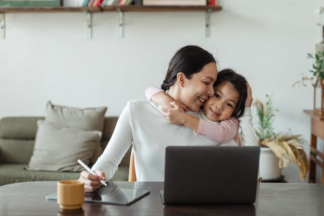 Free Happy young Asian woman working remotely from home with laptop and tablet while adorable little daughter hugging from behind Stock Photo
