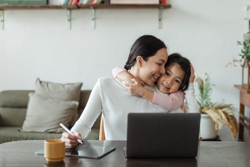 Happy young Asian woman working remotely from home with laptop and tablet while adorable little daughter hugging from behind