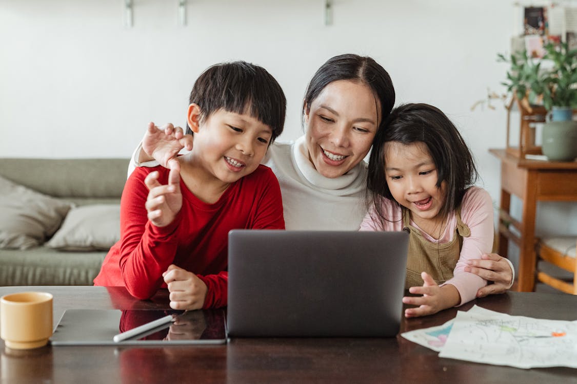 Free Cheerful young Asian woman hugging little kids while watching entertaining program together on laptop during weekend Stock Photo