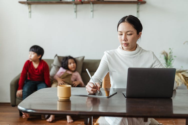 Mum Working On Laptop At Table With Bored Children Behind