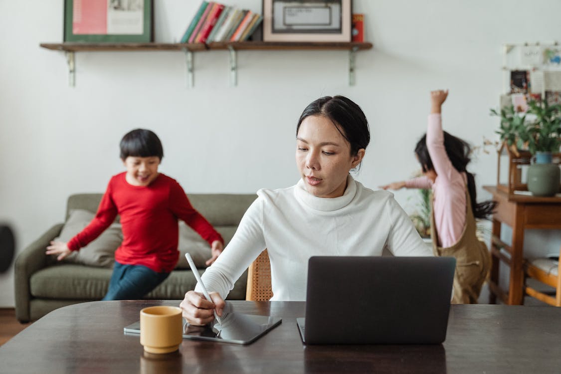 Asian mother working online using laptop and taking notes on tablet with stylus and cheerful children making noise and running behind in living room