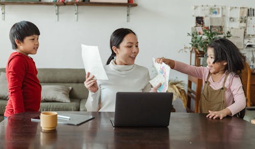 Young Asian mother sitting at table with laptop and tablet in living room and cute children showing mom pictures drawn with crayons