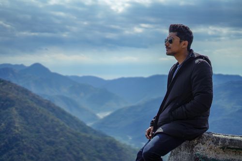 Free Portrait of a man sitting on a mountain overlook, enjoying a serene view with sunglasses and a black jacket. Stock Photo
