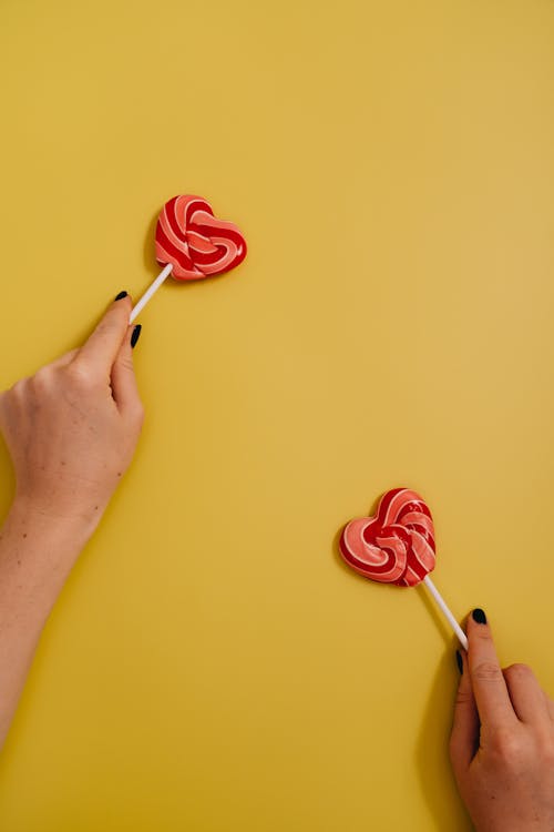 A Person Holding Two Heart Shaped Lollipops Far From Each Other on Yellow Background