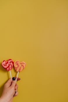 Two heart-shaped lollipops held by a hand against a vibrant yellow background.