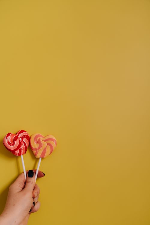 Free Two heart-shaped lollipops held by a hand against a vibrant yellow background. Stock Photo