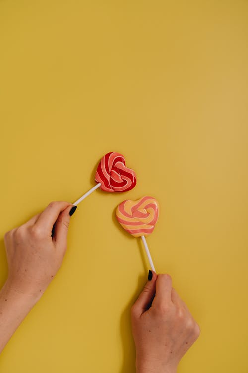 A Person Holding Two Heart Shaped Lollipops Next to Each Other on Yellow Background