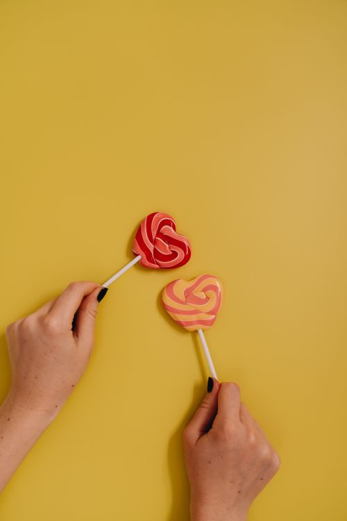 Free Colorful heart-shaped lollipops held against a vibrant yellow backdrop, symbolizing love and sweetness. Stock Photo