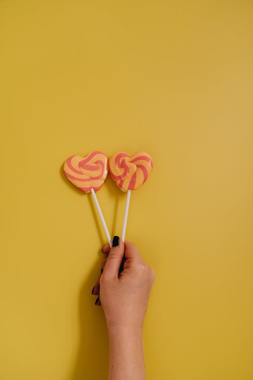 A Person Holding Two Heart Shaped Lollipops Next to Each Other on Yellow Background