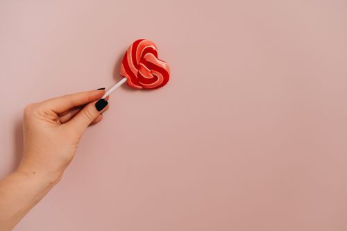 Free A hand holding a heart-shaped lollipop against a pastel pink background, evoking sweet love. Stock Photo