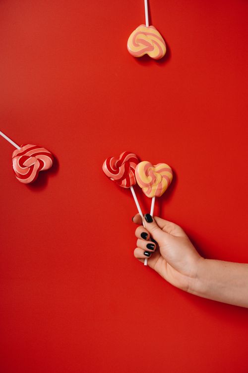 Free Hand holding heart-shaped lollipops against a vibrant red background, evoking sweet love. Stock Photo