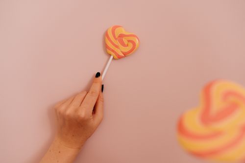 Free A hand with black nails holding a colorful heart-shaped lollipop against a pastel pink background. Stock Photo