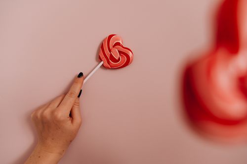 Free A hand holding a heart-shaped lollipop against a light pink background. Stock Photo