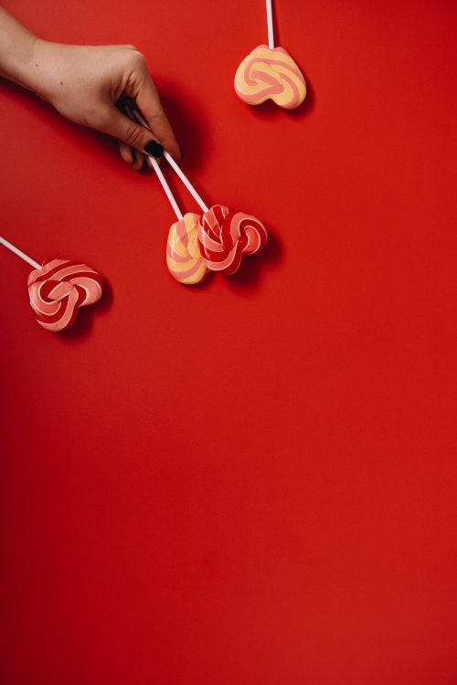 Free Hand holding colorful heart-shaped lollipops against a vibrant red background. Stock Photo