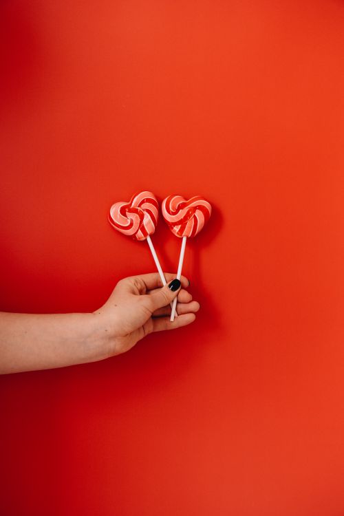Free Hand holding heart-shaped lollipops with a vibrant red background, symbolizing love. Stock Photo