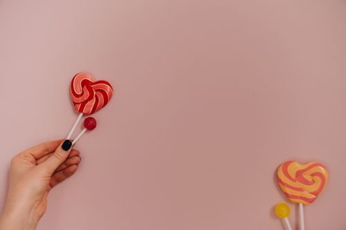 Free Vibrant heart-shaped lollipops held by a hand on a pastel pink background, offering sweet temptation. Stock Photo