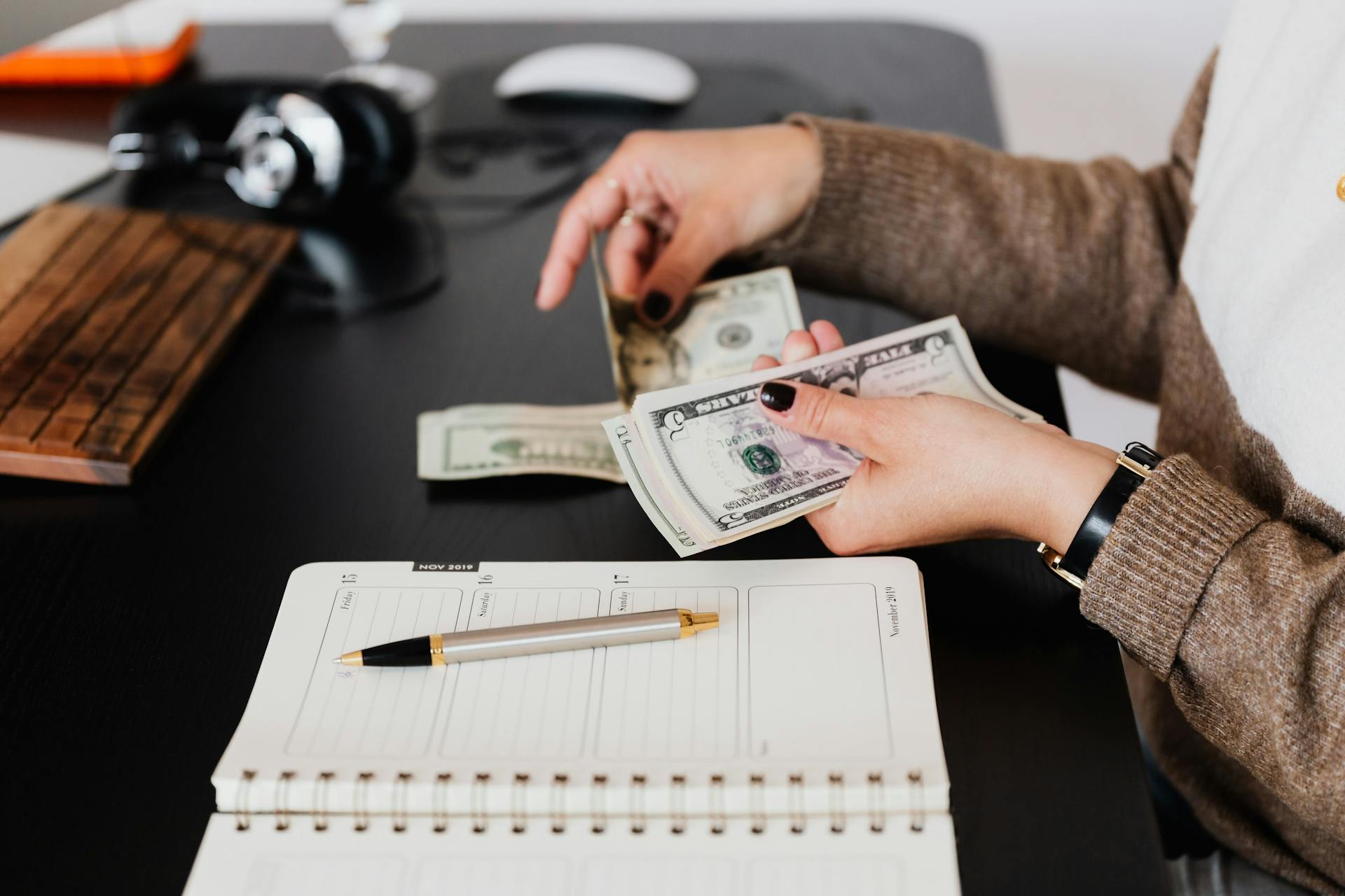 Woman counting cash at desk with notebook, pen, and headphones nearby.