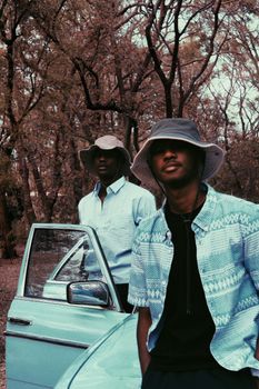 Two fashionable men wearing bucket hats stand by a vintage car in a wooded area.