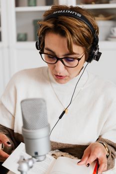 Woman wearing headphones reading aloud from a book into a microphone indoors.