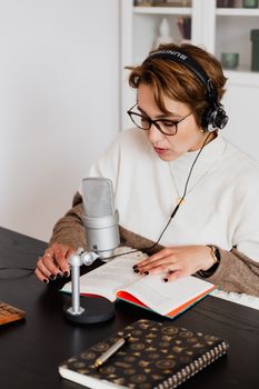 Woman in glasses reading and recording audiobook with a microphone, focused on the literature.