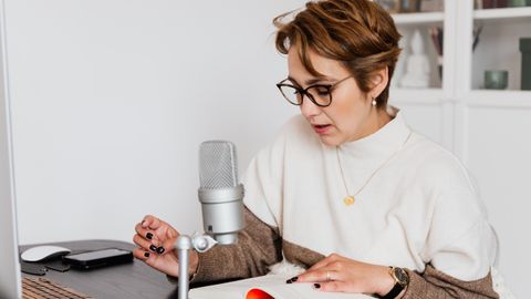 Female narrator in glasses reading out loud from book while sitting at desk with microphone and recording audiobook in cozy study