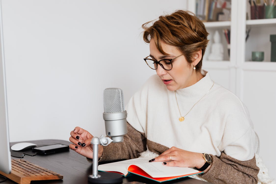 Female narrator in glasses reading out loud from book while sitting at desk with microphone and recording audiobook in cozy study