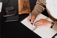 上图中，一位身穿时尚毛衣的女士面目全非地坐在黑色办公桌前，面前配有电脑键盘和智能手机，她一边在记事本上做笔记