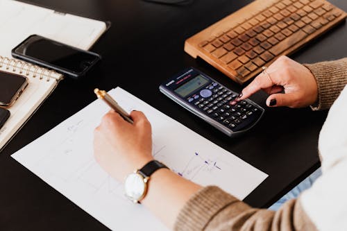 Crop woman using calculator and taking notes on paper