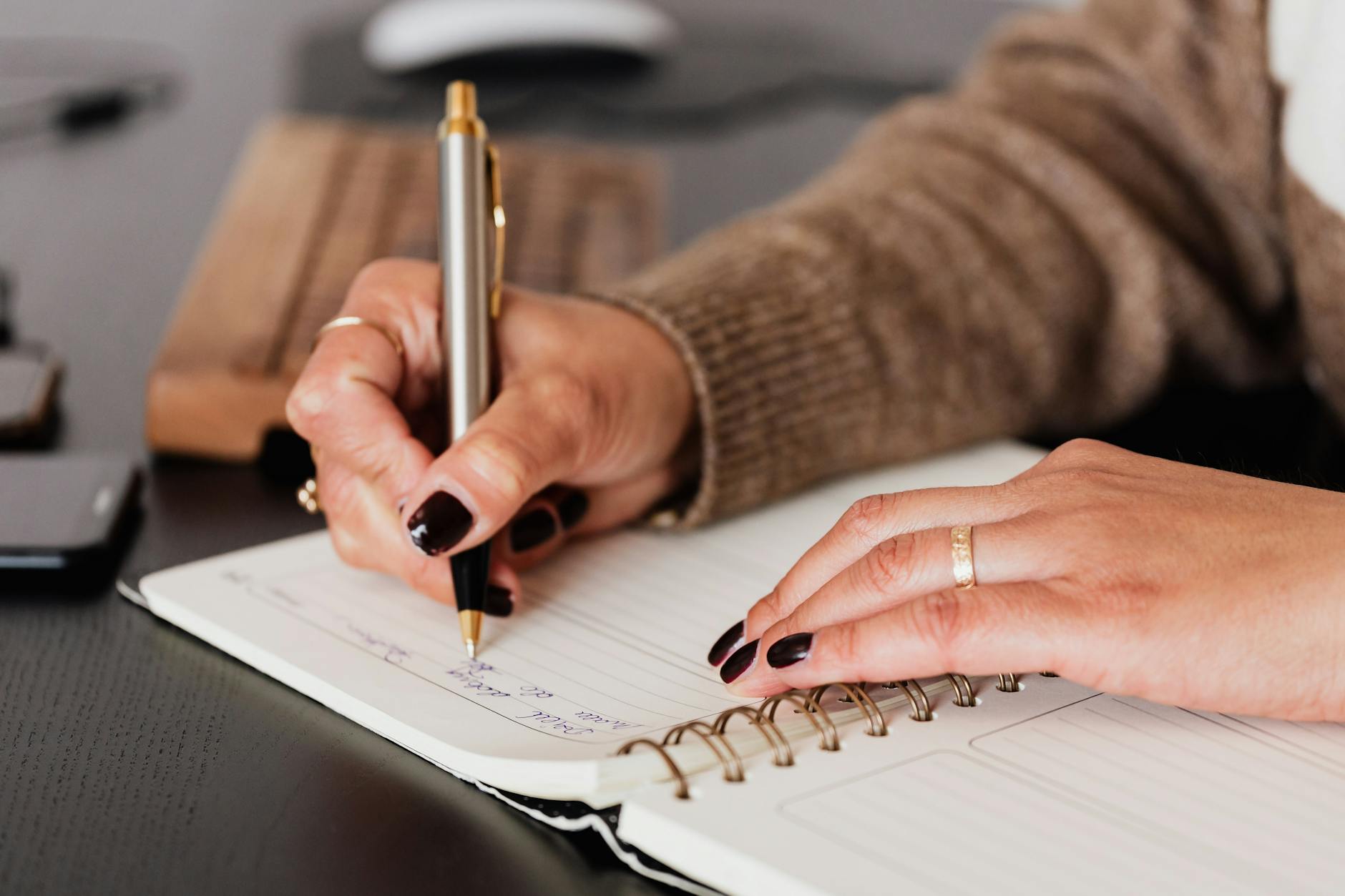 Crop unrecognizable female with stylish manicure sitting at black  desk with keyboard and smartphone and taking notes with silver pen in notepad