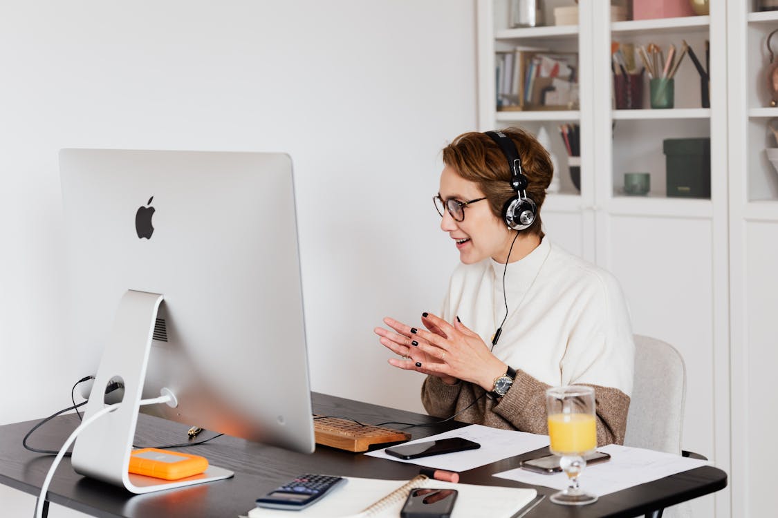 Free Cheerful female in casual wear and wired headphones having conversation via video call on computer while working in light modern workplace Stock Photo