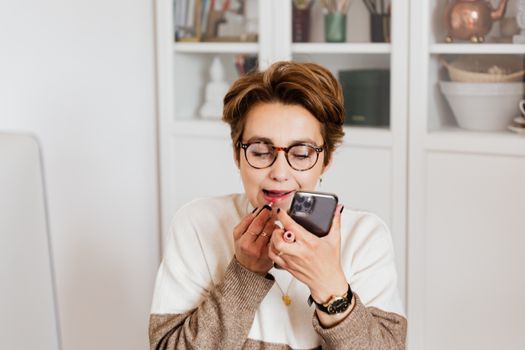 一位女士在室内一边用智能手机，一边涂口红，展现了休闲现代的生活方式